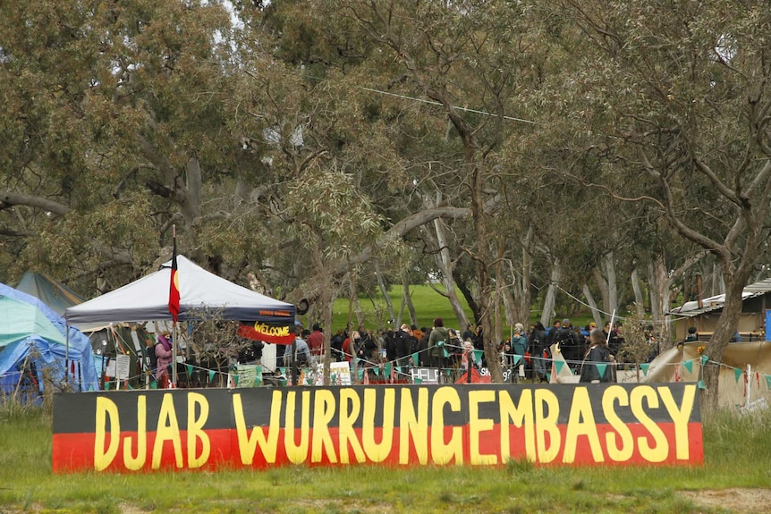 A number of people and tents stand in a bushland setting behind a yellow, red and black sign reading  Djab Wurrung Embassy.