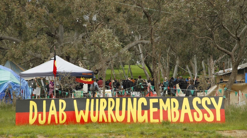 A number of people and tents stand in a bushland setting behind a yellow, red and black sign reading  Djab Wurrung Embassy.