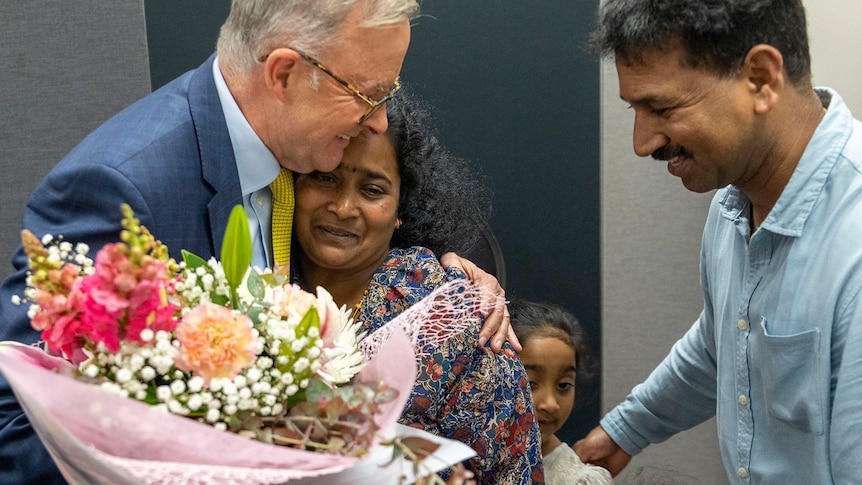 A man with grey hair and glasses in a suit embraces a crying woman as her husband and two daughters look on.