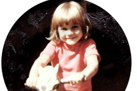 A grainy colour photo of a young girl smiling on a wooden rocking horse.