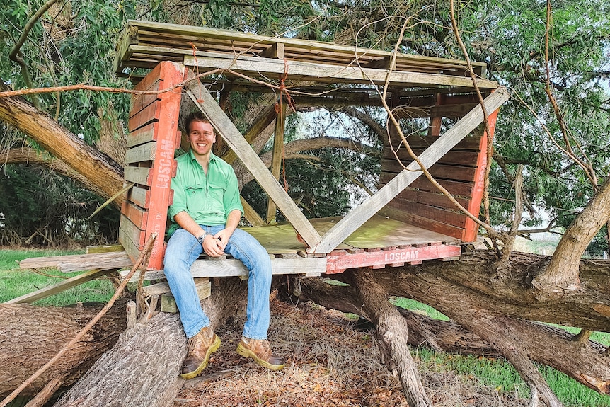 A man in a green shirt, jeans and boots sits in an old wooden treehouse close to the ground.