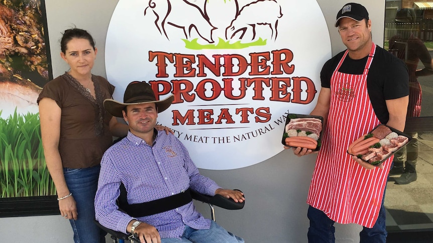 A woman, a man in a wheel chair, and another man holding trays of meat stand in front of a butcher shop