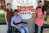 A woman, a man in a wheel chair, and another man holding trays of meat stand in front of a butcher shop