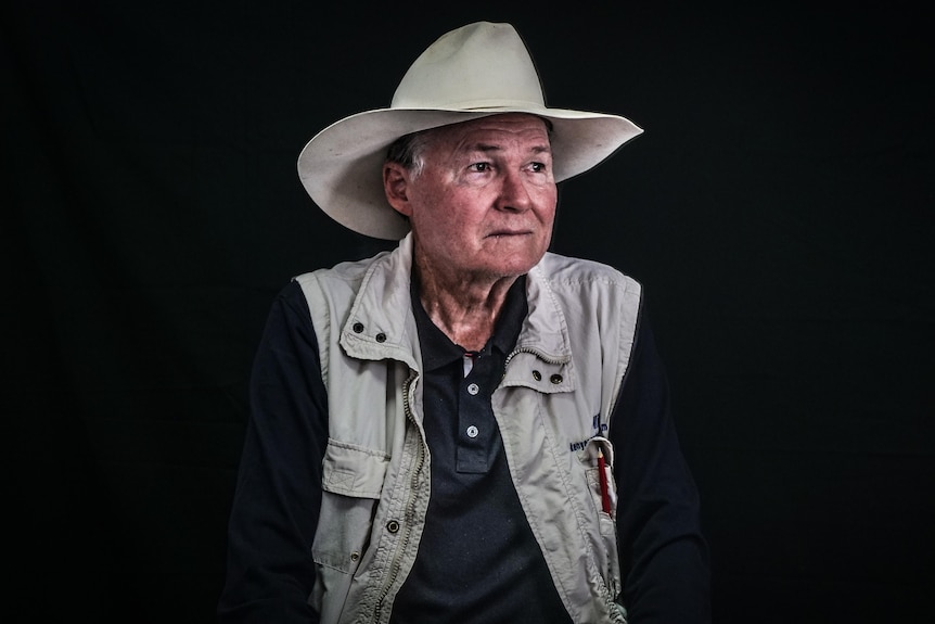 An older man wearing a hat and a hunter's vest posing for a portrait in front of a black background.