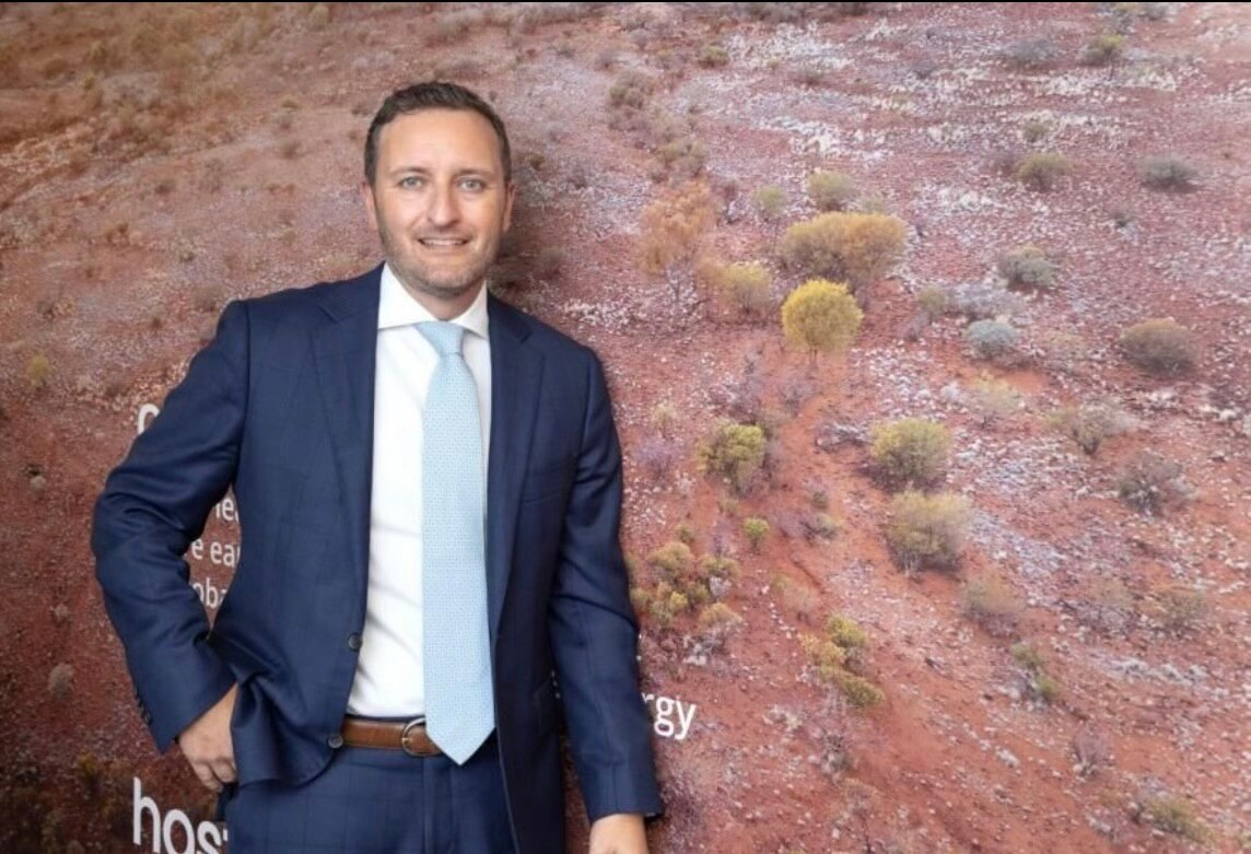 A man in a navy suit stands looking at the camera smiling next to a large landscape photo of Australian shrubs and red dirt land