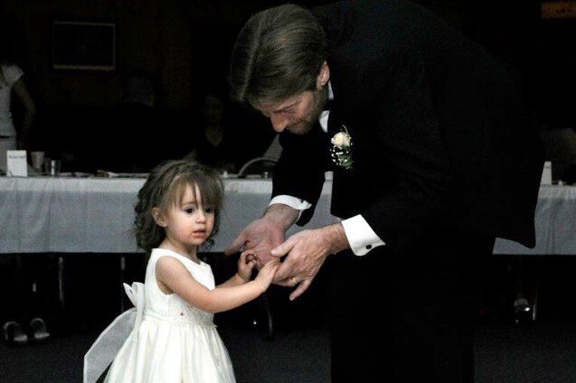 A man in a suit holds hands with a little girl dressed in a white gown