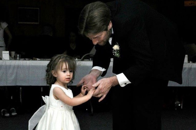 A man in a suit holds hands with a little girl dressed in a white gown