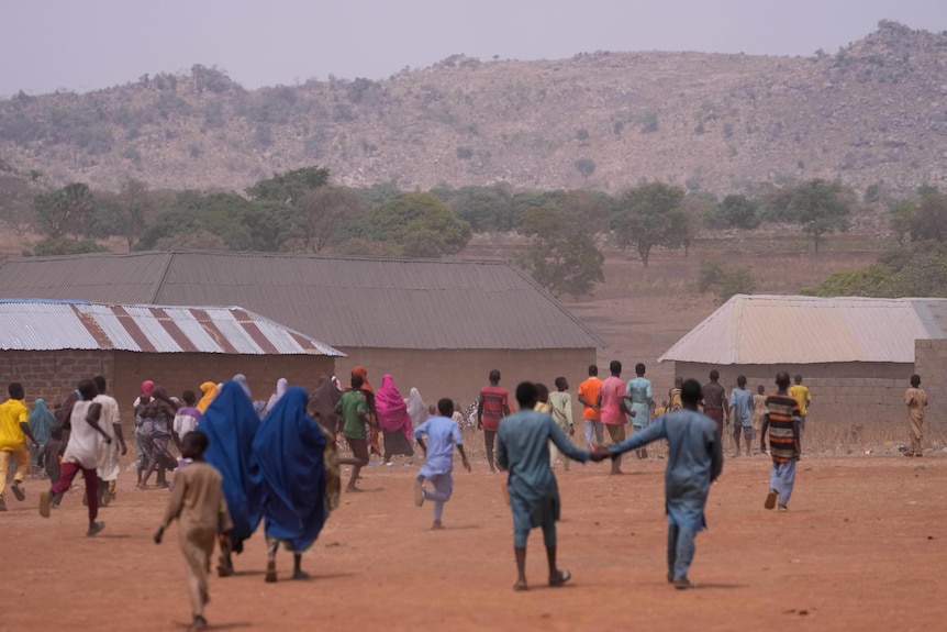 Children and adults in a Nigerian school standing on sandy ground with school buildings in the background.