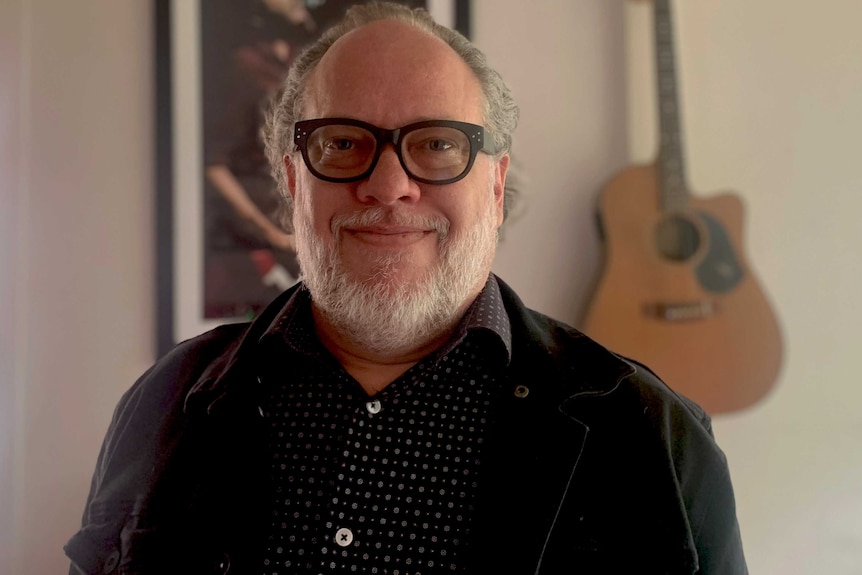 A man with grey hair and thick, black-rimmed glasses stands in front of a wall with a guitar and a framed poster.