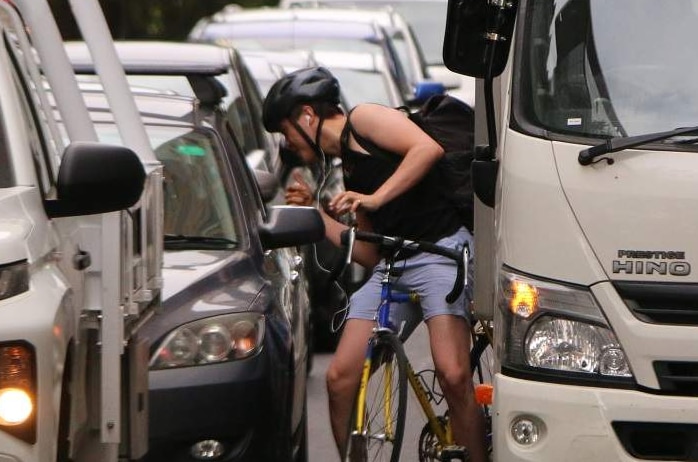A cyclist is wedged in between a car and a parked truck and leans over to look in the window at the driver.