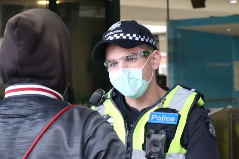 A young man with his back to camera talking to a masked police officer.