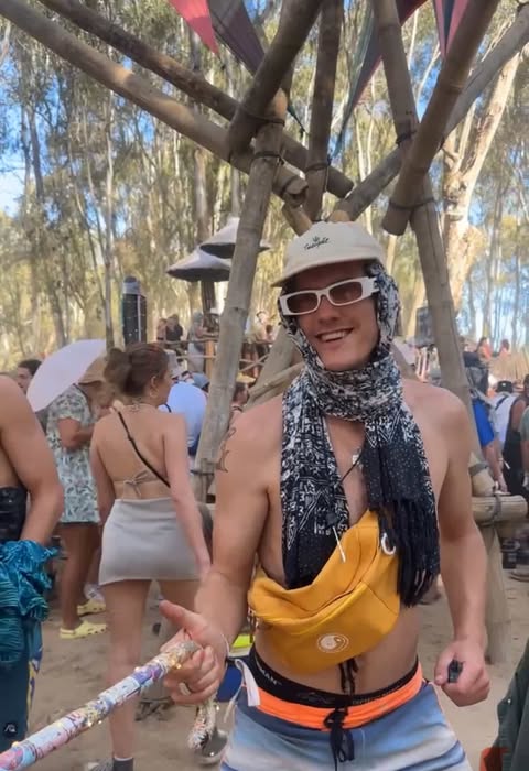 Um jovem em um festival de música segura uma batuta. Ele usa óculos escuros, chapéu e lenço. 