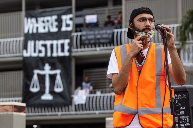 Jonathan Sri lowers a face mask to speak into a microphone as a banner is seen on an apartment block behind him.