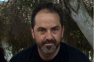 Paul Costa wears a black t-shirt and stands in front of a tree.