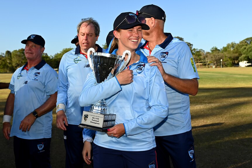 A young white woman smiles as she holds a cricket trophy