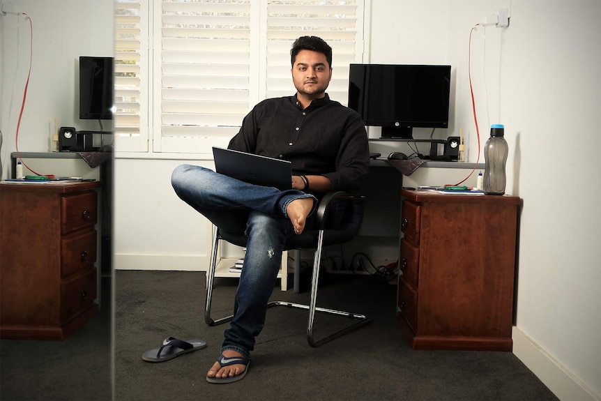 An Indian man wearing a black shirt and a pair of jeans sits in his bedroom at his desk cross legged with laptop in his lap.