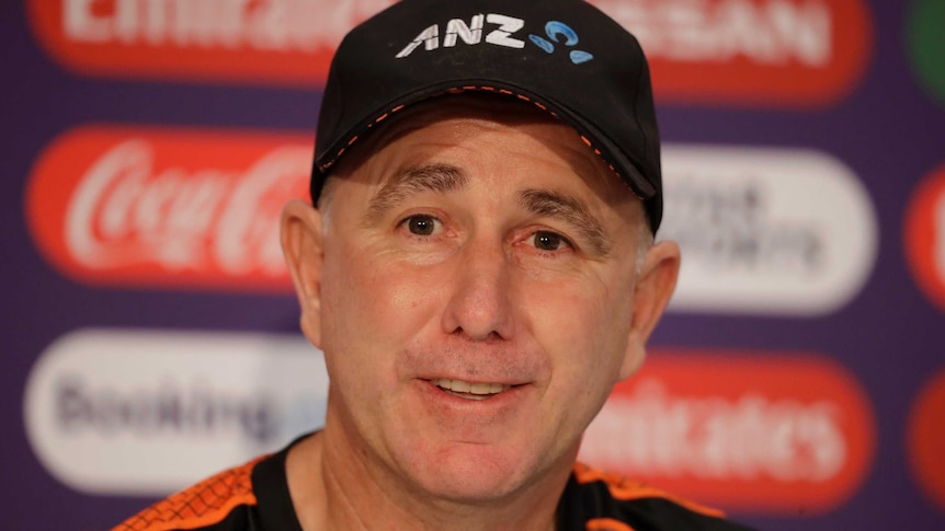 A cricket coach sits at a table to answer questions from the media.