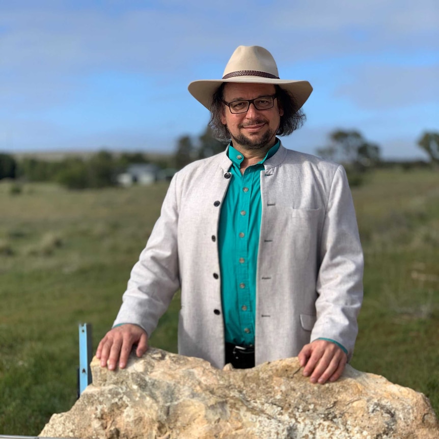 Zuckermann, wearing an turquoise shirt, grey jacket and akubra, stands behind a rock with a street sign for "Nowhere Else Rd"