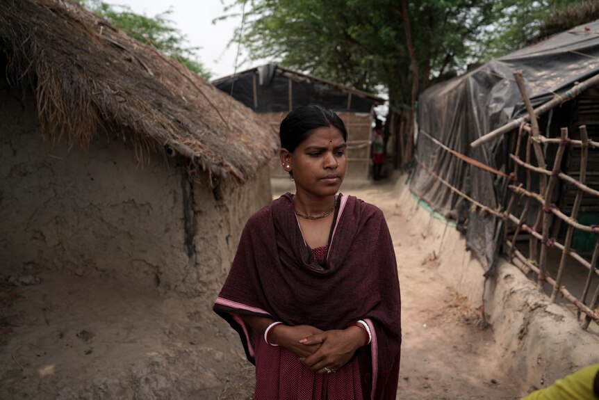 A dark skinned woman wearing a brown sari walks by a house with a straw roof.