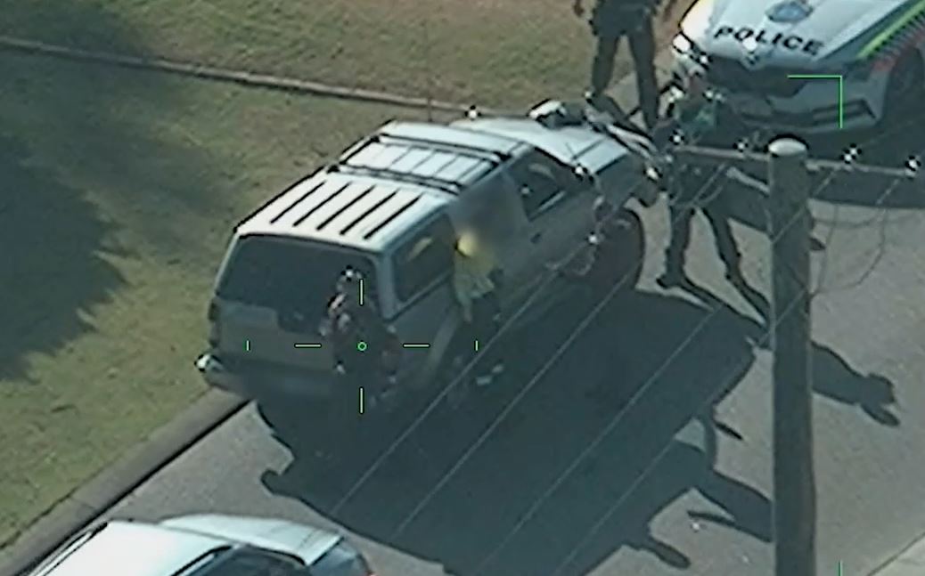 Police surround a man wearing a tradie uniform as he stands next to a silver ute