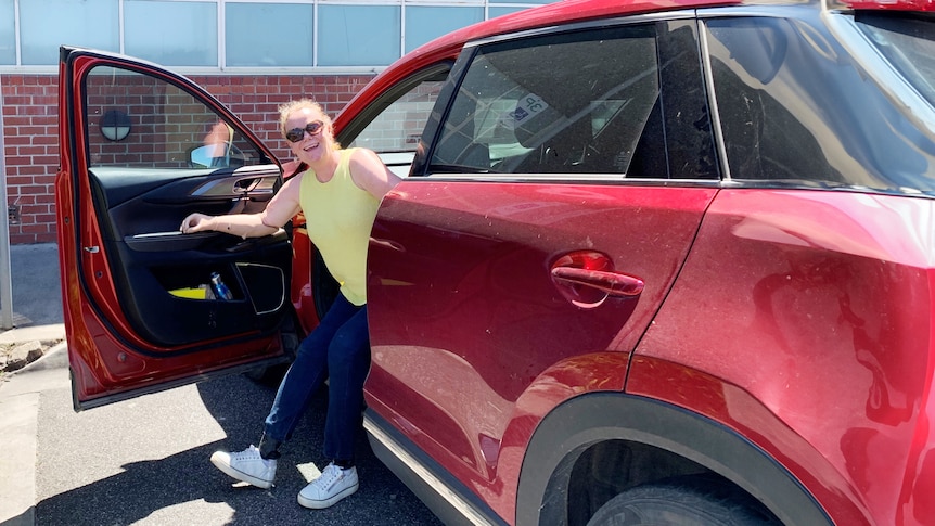 A middle-aged white woman with prosthetic arms and legs exits a red sedan that is parking in an accessible parking bay