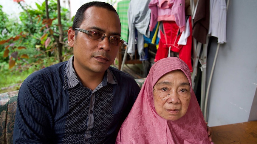 A young Indonesian man and an elderly woman sit together looking pensive while behind them is a garden and washing on a rack.