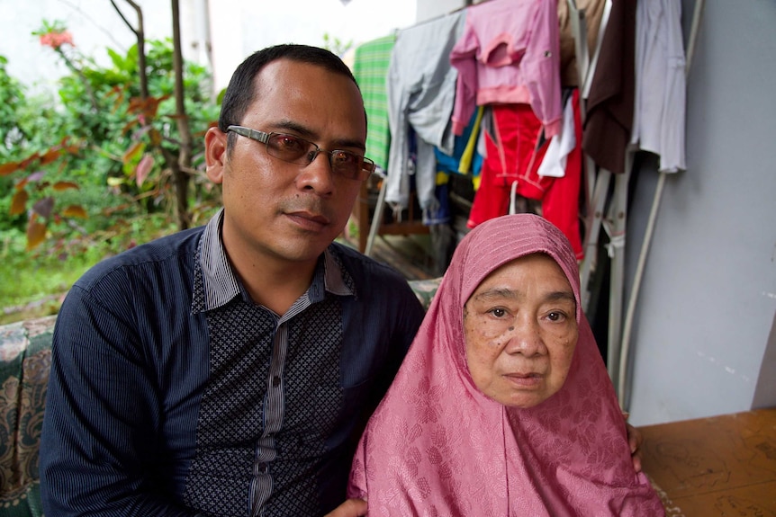 A young Indonesian man and an elderly woman sit together looking pensive while behind them is a garden and washing on a rack.