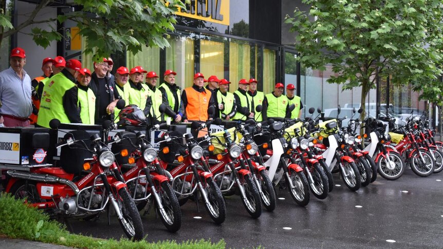 A group of men in red caps and hi-vis vests stand next to their red postie bikes