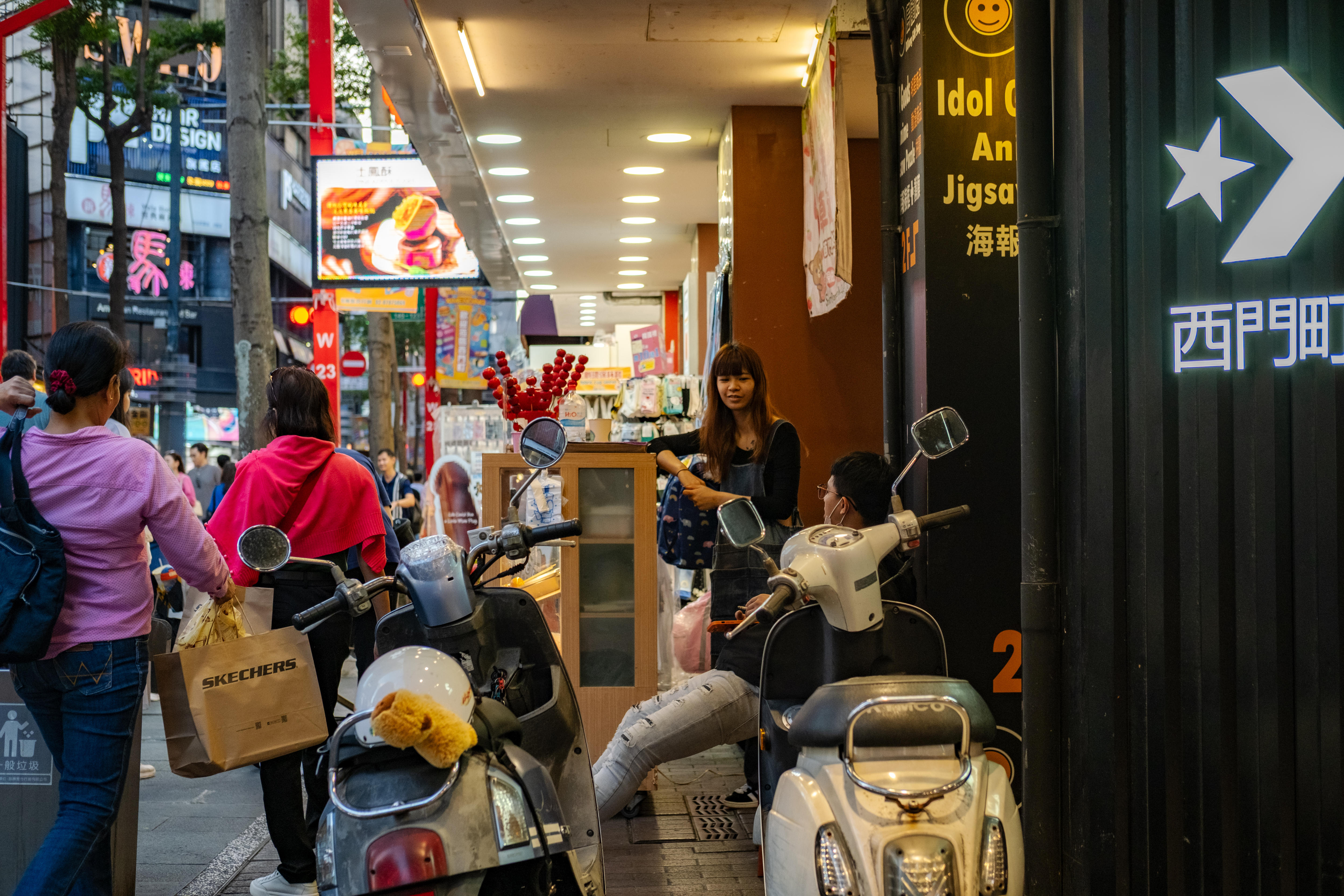 La gente pasa junto a scooters estacionados cerca de la entrada de una tienda.