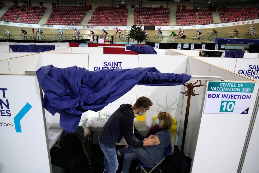 Woman sitting in a folded chair in a white cubical inside a sports stadium getting her COVID-19 vaccine 