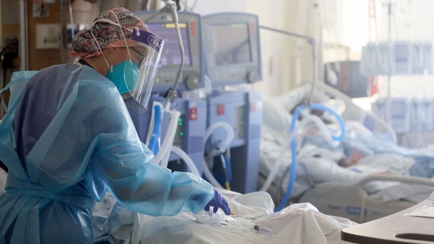 A health worker in full PPE cares for another patient while they are in an ICU.