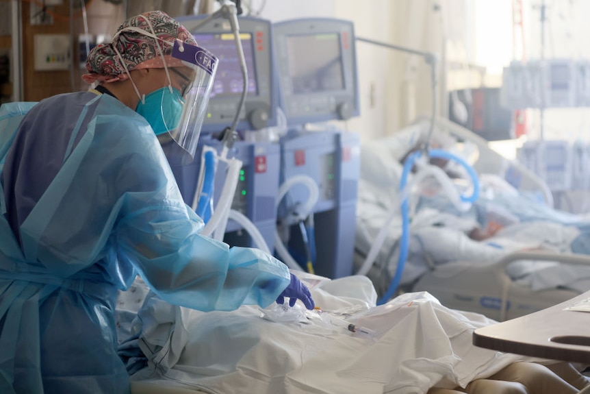 A health worker in full PPE cares for another patient while they are in an ICU.