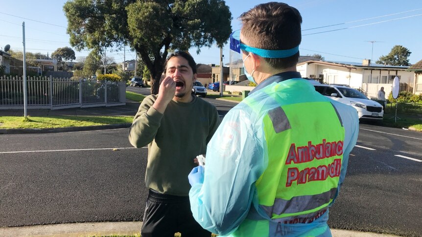 A man wearing a green jumper holds a swab in his mouth while a paramedic in high-vis and PPE stands by.