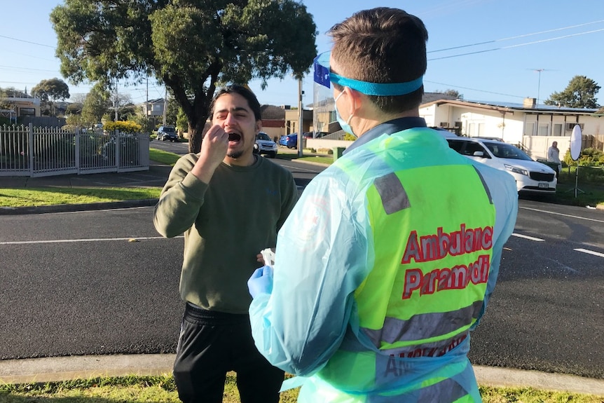 A man wearing a green jumper holds a swab in his mouth while a paramedic in high-vis and PPE stands by.