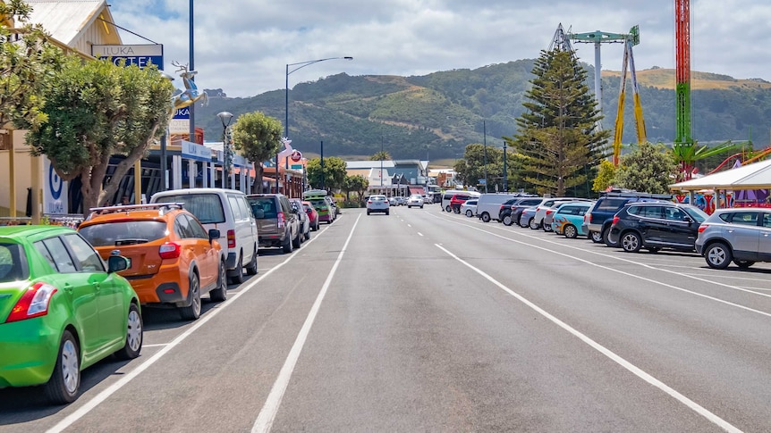 Parked cars on either side of a quiet road through a town with hills in the background