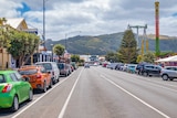 Parked cars on either side of a quiet road through a town with hills in the background