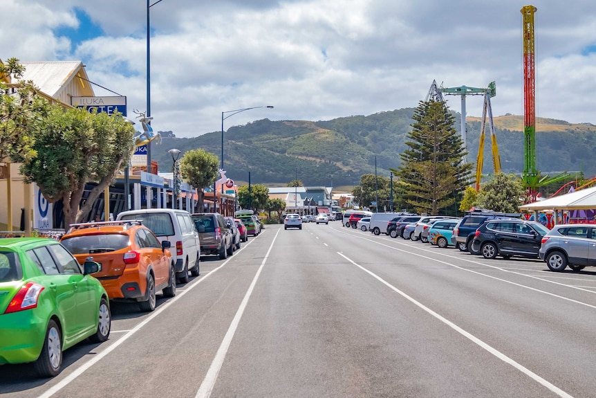 Parked cars on either side of a quiet road through a town with hills in the background