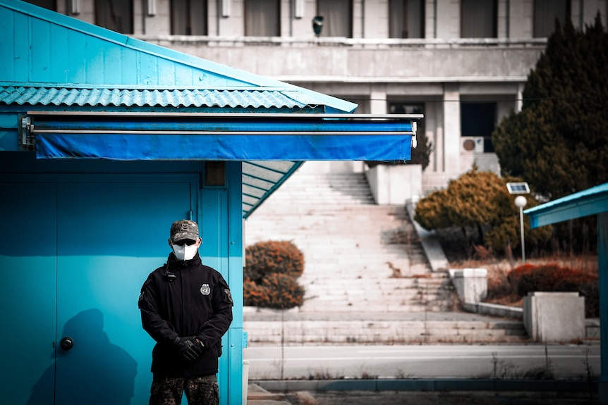Two masked soldiers stand outside two bright blue buildings at the Korean border