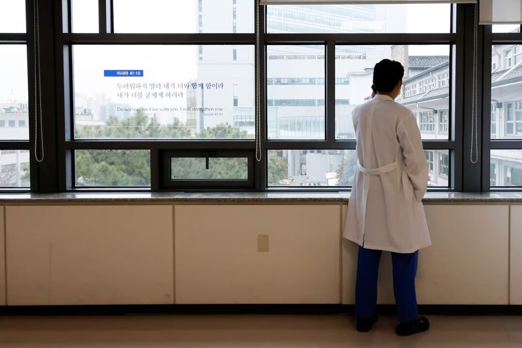 A man in a white coat looks out of a window while on the phone