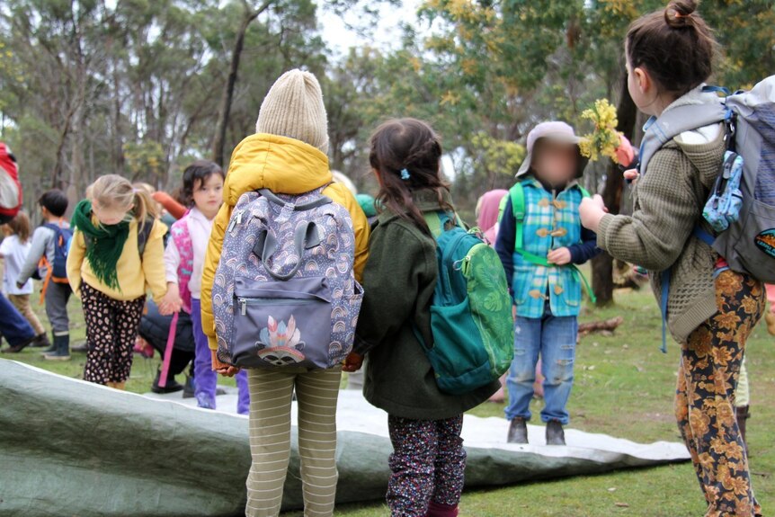 School aged children wearing backpacks as they explore an outside setting, surrounded by tall trees.