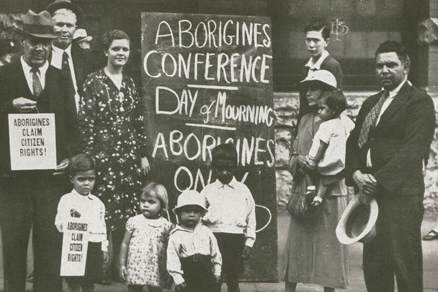 A group of Aboriginal men, women and children in front of protest sign with words day of mourning.