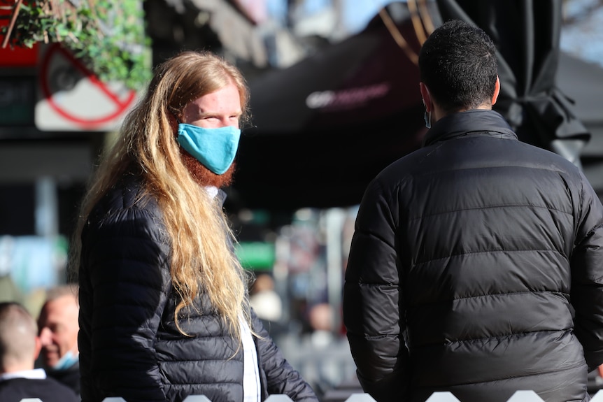 A person with long blonde hair and a red beard wearing a mask on a sunny day.