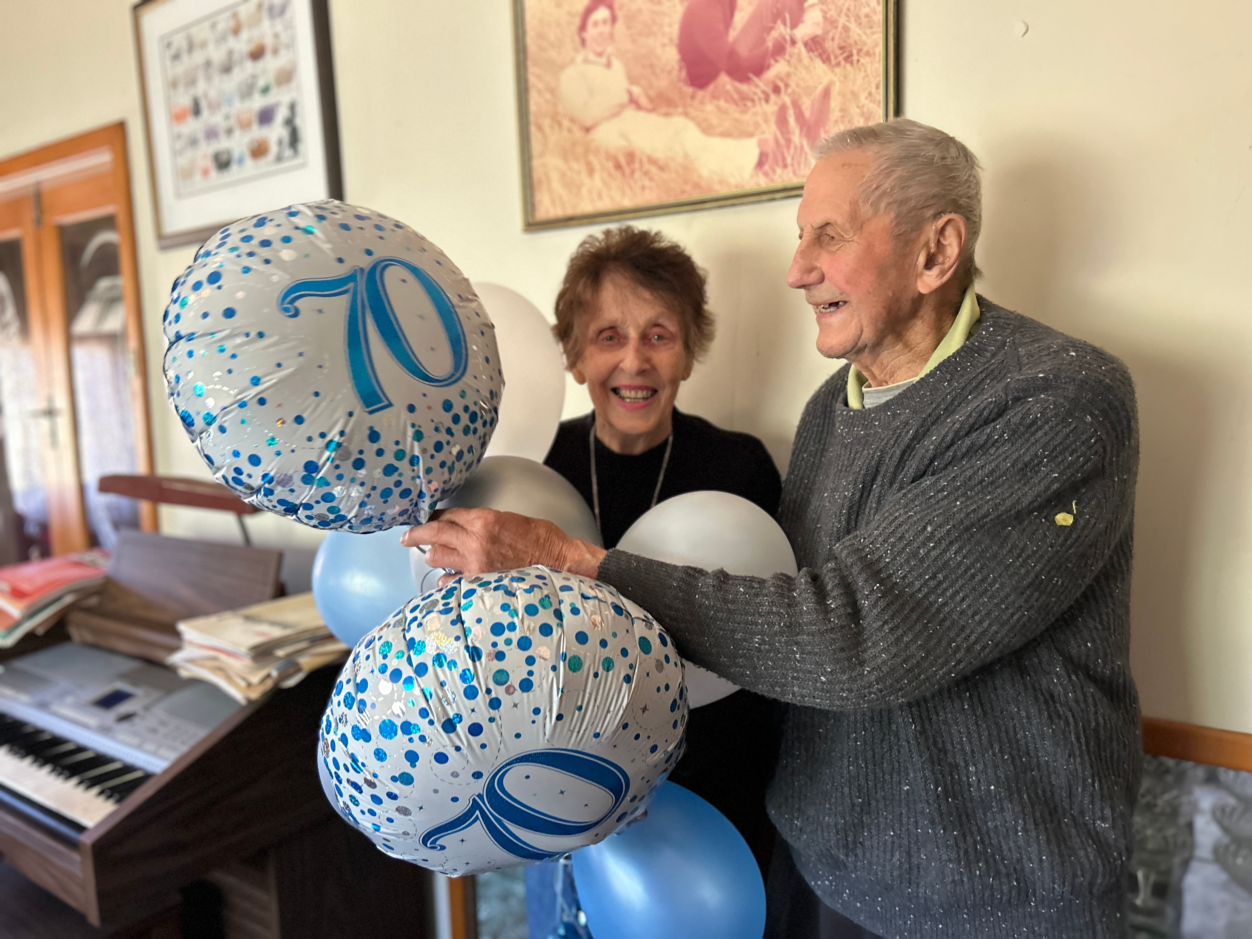 An elderly man and woman smile while holding balloons that say 70.