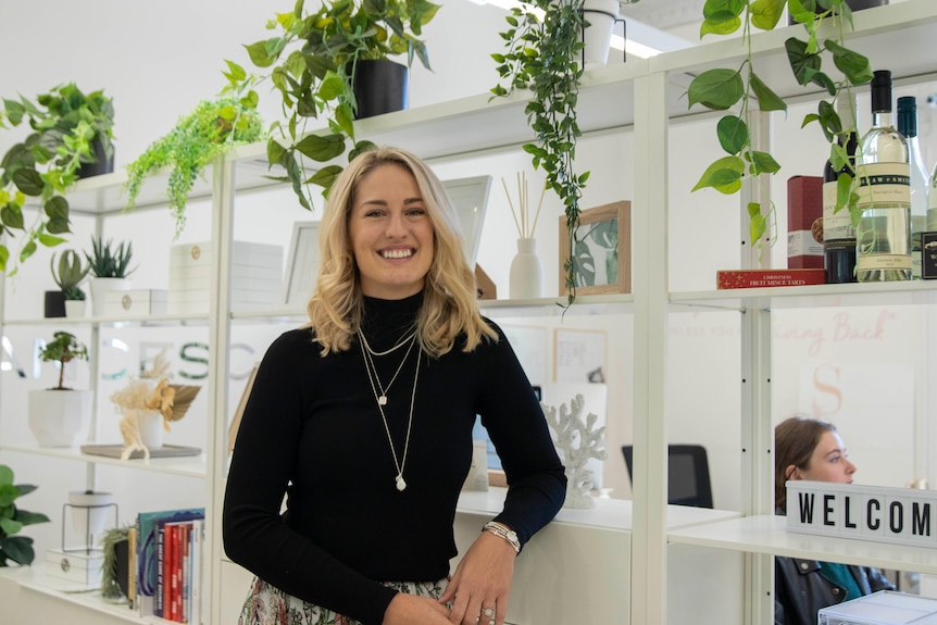 A young blonde haired woman stands in an office with a background of potted plants