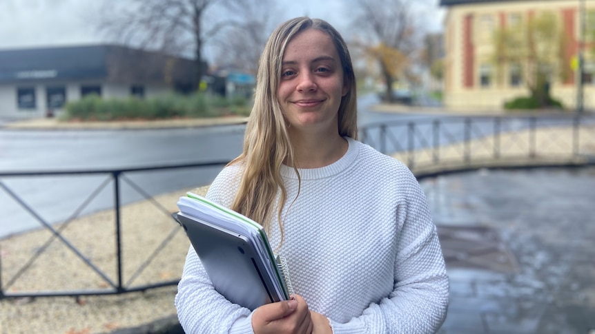 A young woman with long blonde hair stands outside, wearing a white jumper and holding books unde rher arm 