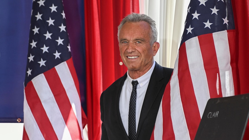 RFK Jr is pictured between two American flags. He looks at the camera and smiles
