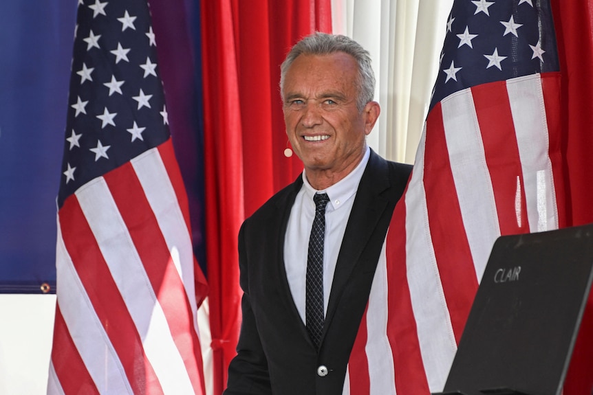 RFK Jr is pictured between two American flags. He looks at the camera and smiles