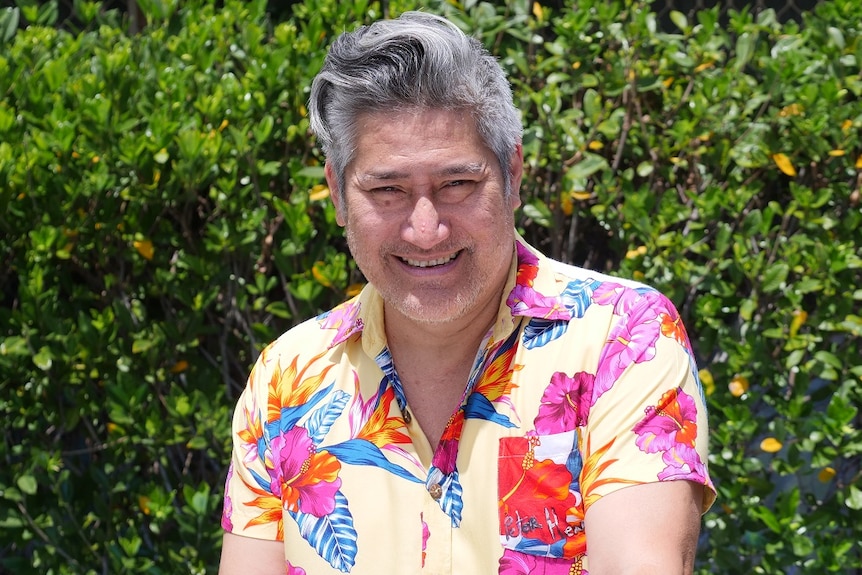 A man with grey hair wearing a yellow, pink and blue coloured shirt smiling in the sun in front of a green hedge