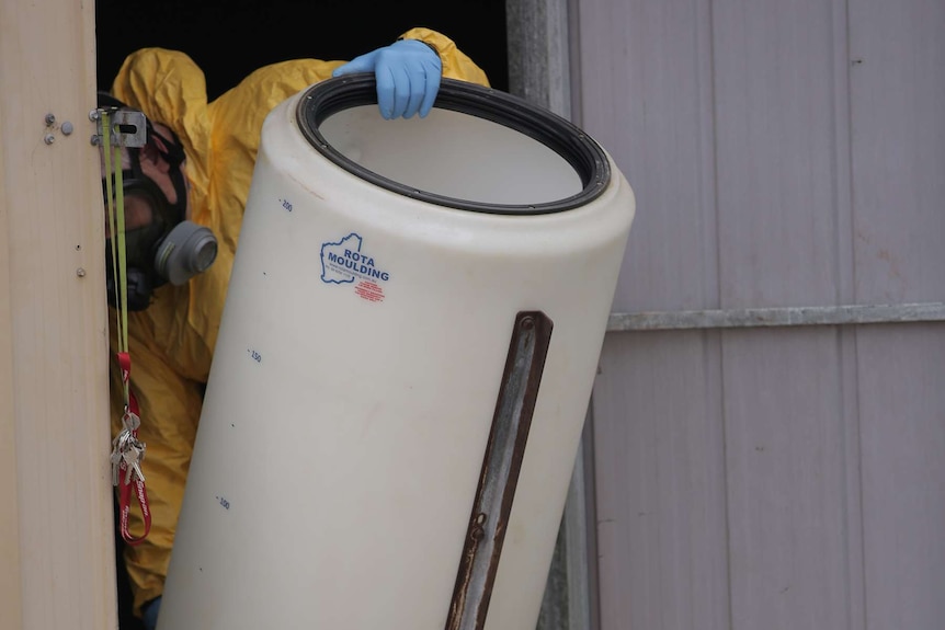 A man in a yellow hazmat suit takes a cannister from a big shed.