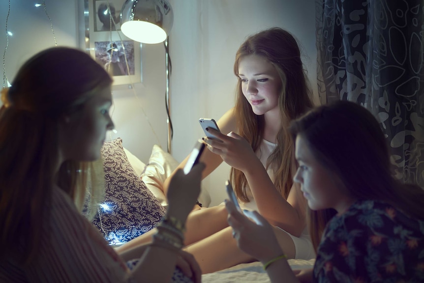 Teenage girls sit in a bedroom looking at their mobile phones.